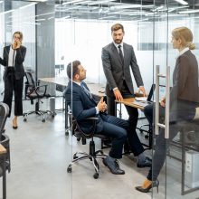 Group of strictly dressed business people having a discussion, sitting at the workplace with a computer in the modern office with glass partitions